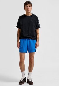 Svart kortärmad skjorta med randiga ärmar, ihop med blå shorts. Outfitten kompletteras av vita strumpor och bruna sneakers.