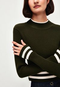 Vihreä ribattu neule, jossa on korkea kaulus ja pitkät hihat, joissa on valkoiset ja mustat raidalliset hihansuut. Lyhennetty malli, jossa on teksturoitu kangas.