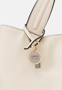 Kremowa torebka z imitacji skóry z fakturowanym wykończeniem, ozdobiona czarną lamówką, podwójnymi uchwytami oraz złotym zawieszką z wygrawerowanym napisem "GUESS EST. 1981".