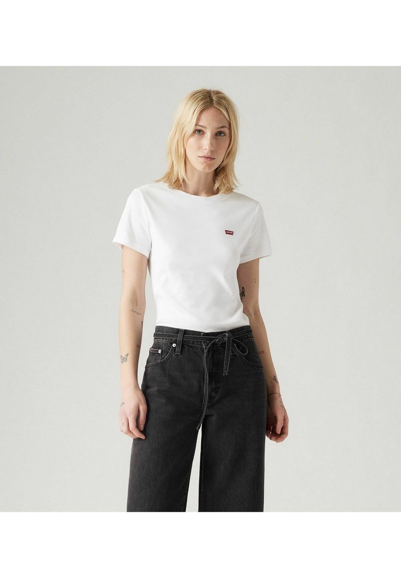 Camiseta de algodón blanca con cuello redondo y mangas cortas, que presenta un pequeño logo rojo en el lado izquierdo. Combinada con jeans negros de tiro alto.