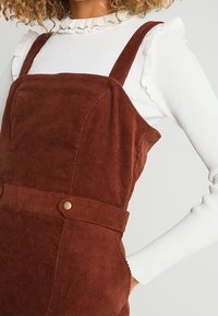 Hnědé kordové lacláče se širokými ramínky, s ozdobným knoflíkem v pase, doplněné bílým pruhovaným topem s dlouhým rukávem a řaseným výstřihem.