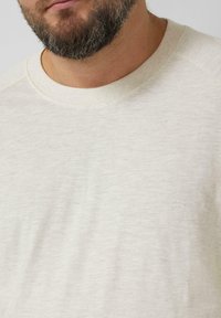 Jasnobeżowa koszulka z okrągłym dekoltem, wykonana z teksturowanej tkaniny. Posiada prążkowany dekolt i rękawy typu raglan. Gładka powierzchnia, minimalistyczne elementy designu.