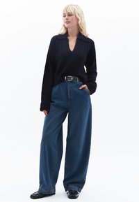 Maglione blu navy a costine con scollo a V, abbinato a jeans blu a gamba larga a vita alta. Cintura nera con accessori in argento, scarpe nere.