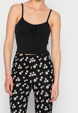 Kobieta w czarnym krótkim topie na ramiączkach i czarnych spodniach w biały wzór stokrotek.