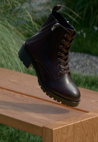 Stivale alla caviglia in pelle marrone scuro con lacci e zip laterale, appoggiato sul bordo di una panchina di legno all'aperto con erba sullo sfondo.