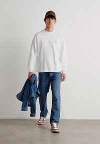 Camisa blanca de manga larga, jeans azules, sosteniendo una chaqueta de mezclilla. Gorra marrón y zapatillas con acentos en blanco, marrón y beige. Fondo minimalista.