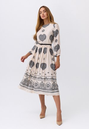 Kobieta ubrana w kremową sukienkę do kolan z czarnymi, misternymi wzorami kwiatów i serc, z długimi rękawami, czarnym paskiem oraz beżowymi szpilkami, stoi na białym tle.