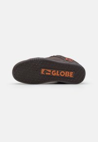 Tamno smeđa guma na đonu tenisica sa teksturiranim gripom, sa izraženim "GLOBE" logotipom u narančastoj boji i uzorkom malih krugova i linija.