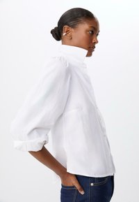 Chemise blanche à boutonner, avec un col haut et des manches bouffantes. Confectionnée en matériau léger, elle présente une coupe décontractée et des poignets retroussés.