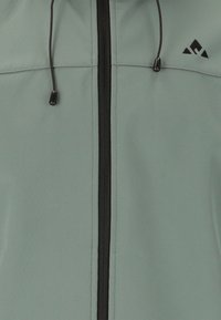 Veste soft-shell vert clair avec fermeture éclair noire et cordon de serrage. Présente un petit logo noir sur le haut à gauche. Texture lisse et design fonctionnel.