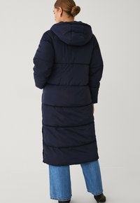 Marineblauwe pufferjas met een capuchon, met een lange, gestructureerde vorm en horizontale stiksels. Draagt over blauwe denimbroeken en zwart leren laarzen.