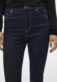 Tamno plave traperice s uskim krojem. Značajke uključuju žute šavove, prednje džepove i pojas s kopčanjem na dugme.