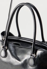 Κοντινή λήψη μαύρης δερμάτινης τσάντας χειρός με διπλές στρογγυλές λαβές και ασημένιους μεταλλικούς κρίκους σε ανοιχτό φόντο.