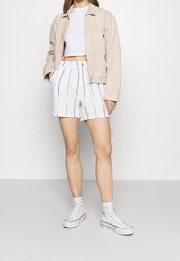 Femme portant une veste beige, un crop top blanc, un short blanc à rayures et des baskets montantes blanches, debout devant un fond uni.