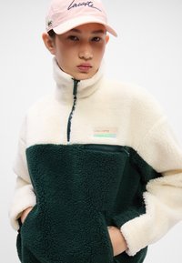 Jakna s polu-zipom od flisa s bojama krem i tamnozelenu, prednji džep i Lacoste logotip. Nosite s svijetloružičastom kapom.