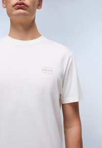 Tricou alb cu mâneci scurte. Prezintă un mic emblema a steagului, discret, pe pieptul stâng. Este fabricat dintr-un material neted și ușor.