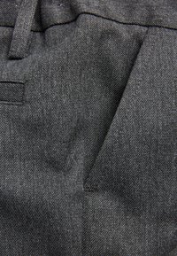 Tissu gris présentant un tissage texturé, avec un détail de poche visible et des coutures piquées. Le matériau semble résistant et de poids lourd.