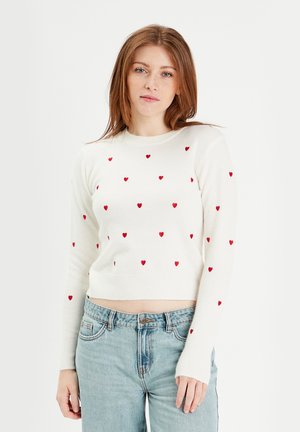 Maglione crema a maniche lunghe con motivo a cuori rossi. Design cropped con collo e polsini a costine. Indossato con jeans a vita alta.