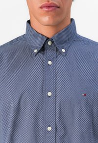 Hombre con camisa de botones azul de patrones, con pequeños rombos blancos y marinos y botones blancos, logo parcialmente visible en el pecho.