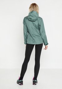 Lett, hættejakke i dæmpet petrol med en glat, skinnende overflade, parret med sorte leggings og multibevægelsessko til vandring.