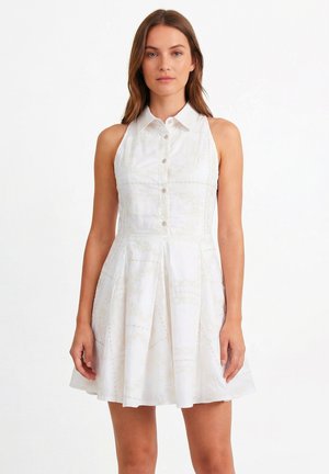 Kobieta ubrana w białą sukienkę bez rękawów z kołnierzykiem, guzikami z przodu i rozkloszowaną spódnicą, stojąca na białym tle.