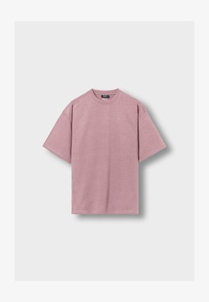 Prosty, luźny, krótki t-shirt w kolorze bladoróżowym z okrągłym dekoltem, wyłożony na płasko na jasnym tle.