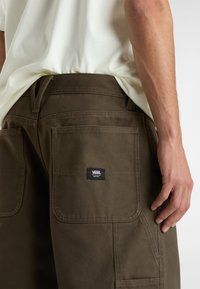 Braune Baumwoll-Cargohosen mit mehreren Taschen, einem Rückenteil mit dem Vans-Logo und einem sichtbaren Taillenbund. Kombiniert mit einem hellen Hemd.
