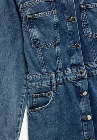 Denim-hame keskisinisessä, jossa on napitus edessä, kaksi rintataskua ja vyötäröllä vyö. Ominaisuuksiin kuuluu kontrastivärinen ommel ja metallinapit.