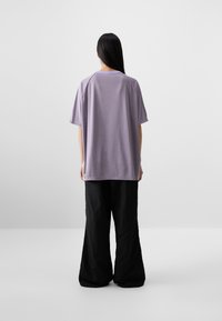 Una persona sta in piedi di spalle, indossando una camicia larghi color lavanda e pantaloni ampi neri su uno sfondo grigio uniforme.