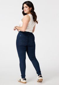 Donkerblauwe skinny jeans met een hoge tailleband, vijf zakken en een gladde denimtextuur. Gecombineerd met een aansluitend wit tanktop en sneakers.