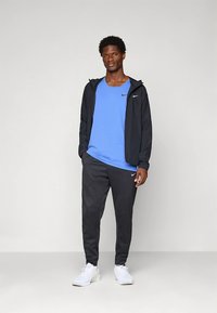 Herrensportbekleidung mit blauem Tanktop, schwarzer Jacke mit Reißverschluss und schwarzen Jogginghosen. Das Material wirkt leicht und hat eine glatte Textur.