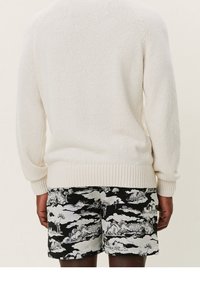 Stickad krämfärgad tröja med ribbad nederkant, kombinerad med svarta shorts med ett vitt molnmönster och landskapsgrafik.