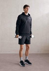 Sort hoodie med et logo, sorte shorts, grå sokker og sorte sneakers. Personen holder sølv håndvægte i en neutral stilling.