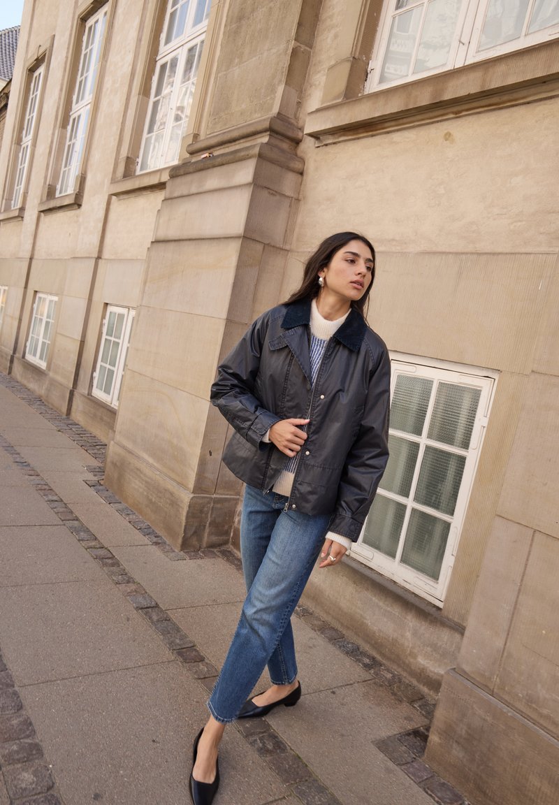 Schwarze, glänzende, leichte Jacke mit einem Kragen, kombiniert mit einem weißen Pullover und blauen Jeans. Das Model steht auf einem Steinpflaster nahe einem Gebäude.