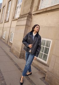 Svart, glänsande, lätt jacka med krage, tillsammans med en vit tröja och blå jeans. Modellen står på en stenlagd gångväg nära en byggnad.