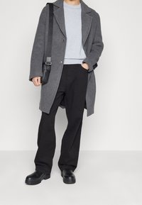 Grå ullrock med snedskurna slag, svart knappstängning, bärs över en ljusgrå tröja och svarta byxor, ihopparad med svarta plattformsskor.