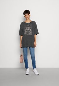 T-shirt oversized de cor carvão com estampa gráfica, mangas curtas, emparelhado com jeans azul claro e ténis brancos, segurando uma mala rosa.