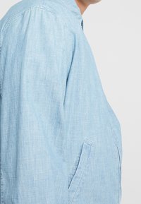 Ljusblå lätt chambray-jacka med ståkrage, sidofickor och avslappnad passform. Tyget har en subtil textur.