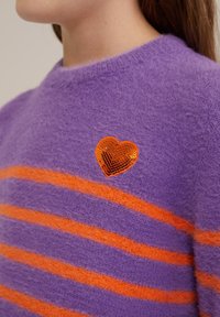 Vijolična pulover s pomarančastimi vodoravnimi črtami, izdelan iz puhastega materiala. Ima pomarančno sekvinskko srčno nalepko na prsih.