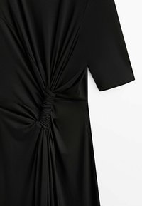 Robe noire avec manche mi-longue présentant un détail de nœud plissé à la taille, créant des plis de tissu rassemblés.