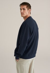Man in een marineblauwe gestructureerde trui en beige broek, zijwaarts staand met de handen in de zakken tegen een effen achtergrond.