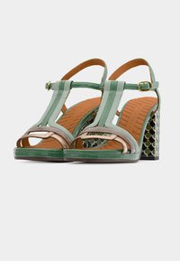 Πράσινα σανδάλια από δερματίνη με σχεδιασμό T-λάστιχου και χρυσές λεπτομέρειες. Το τακούνι είναι χοντρό και έχει υφή με σχέδιο.