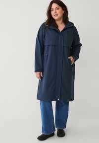 Marineblauwe waterdichte jas met een capuchon, twee voorzakken en een met rits afsluitbare borstzak. Gecombineerd met spijkerbroek en zwarte instapschoenen.