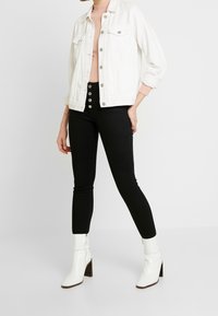 Chaqueta de mezclilla blanca sobre un suéter rosa, combinada con jeans negros de cintura alta con botones plateados y botines blancos de tacón.