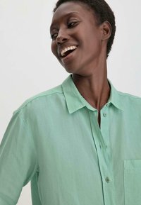 Helle grüne Hemd mit Knöpfen aus einem weichen, leichten Stoff; verfügt über einen Kragen und eine einzelne Brusttasche. Glatte Textur mit subtiler Ausstrahlung.