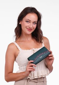 Teal leather wallet with wave pattern, purple accent and polka dots. Features fold-over closure. Model holds wallet against a white background.