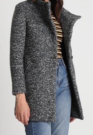 Femme portant un manteau croisé texturé gris foncé sur un haut rayé jaune, noir et blanc, associé à un jean bleu.