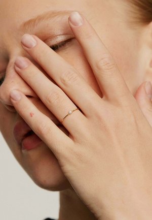 Anillo de oro con un pequeño acento de diamante, llevado en una mano. La mano cubre parcialmente una cara, mostrando un esmalte de uñas natural y la textura de la piel.
