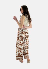 Witte broek met een bruin loofpatroon, elastische tailleband en wijde pijpen, gecombineerd met een aansluitende beige top en lichte schoenen.