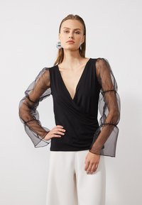 Svart innpakningstopp med dyp V-hals og gjennomsiktige, puffete svarte organza-ermer. Kombinert med hvite vidde bukser. Glede stoff med figursydd silhuett.